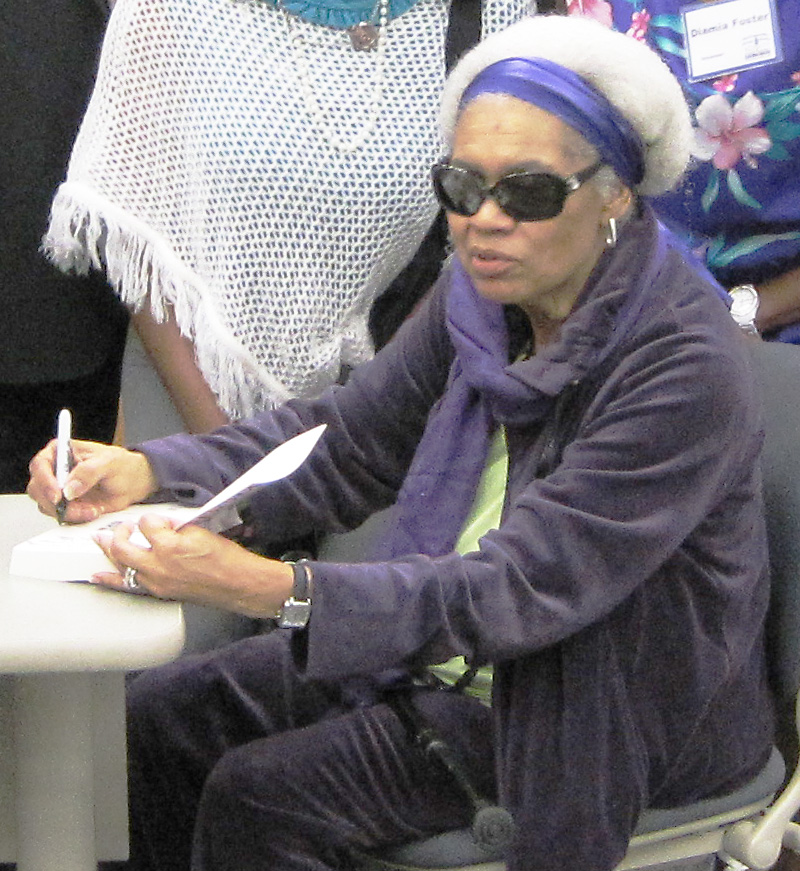 Due sits at a table, holding a pen and signing a book. She is looking up and focusing beyond the camera.