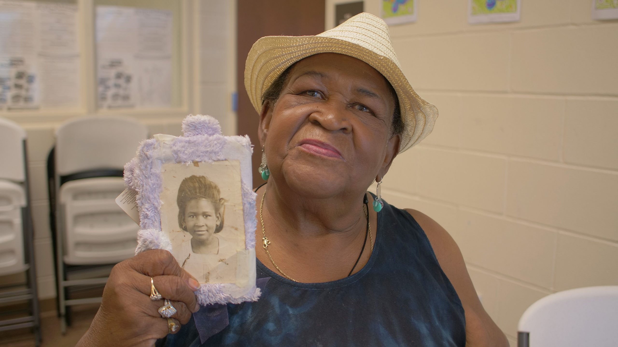 Marshall holds a framed photo of herself as a child. She is sitting in a folding chair and looking at the camera.