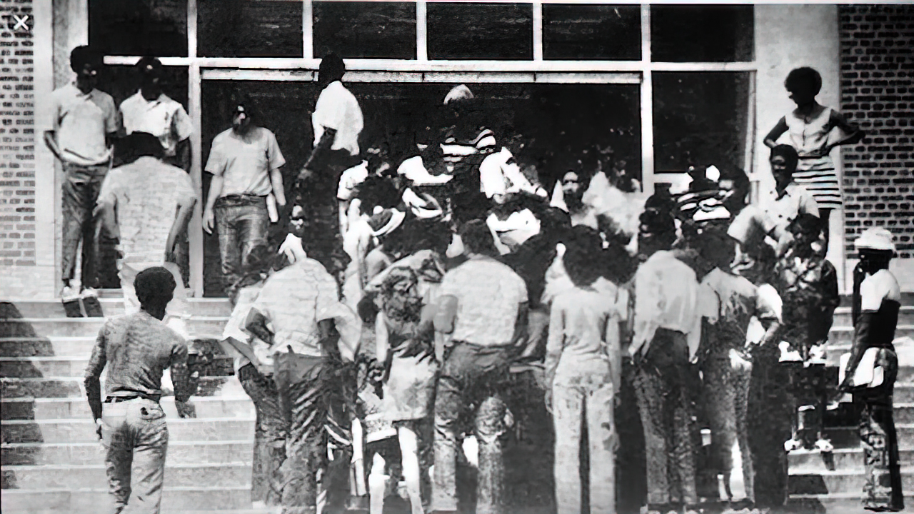 Black and white photo of students standing in a circle on the front steps to Tigert Hall. Most face away from the camera, toward the building doors.