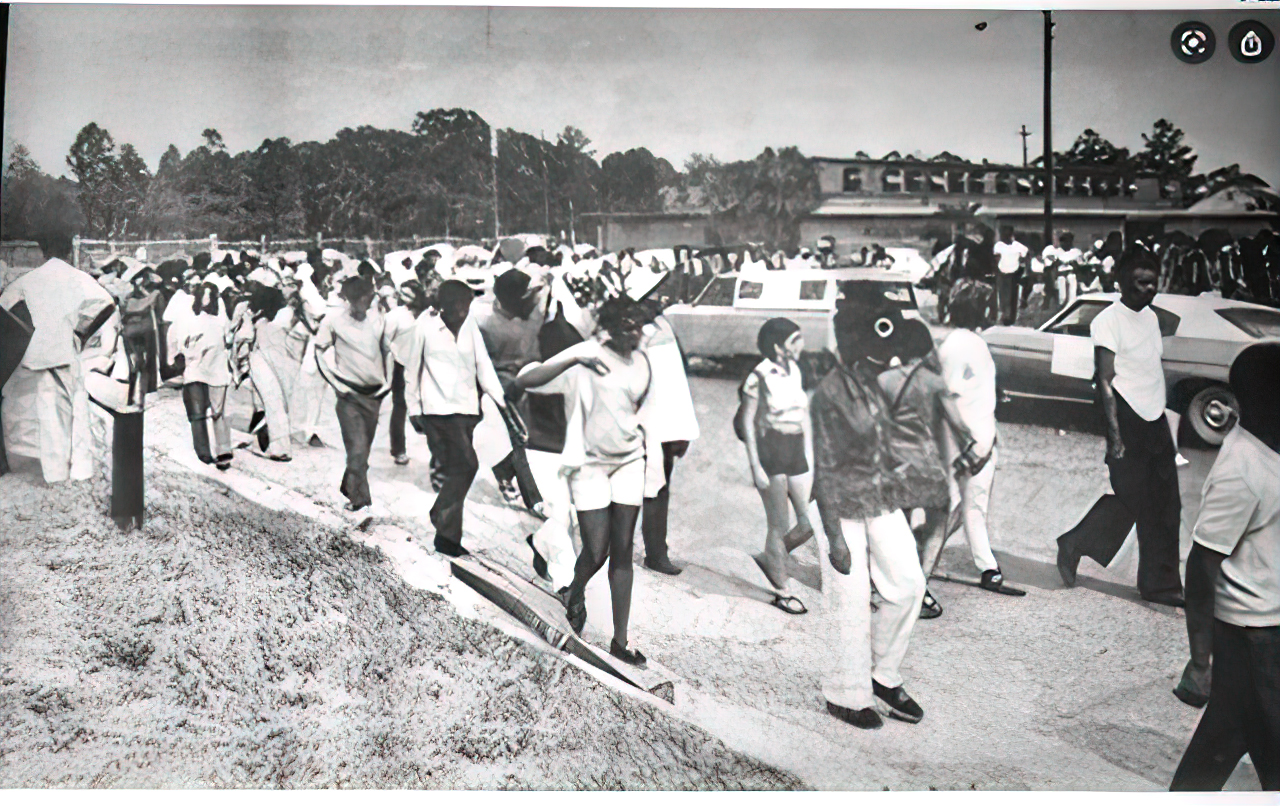 Black and white photo of a group of students marching along two sides of the sidewalk as cars drive down the street between them.