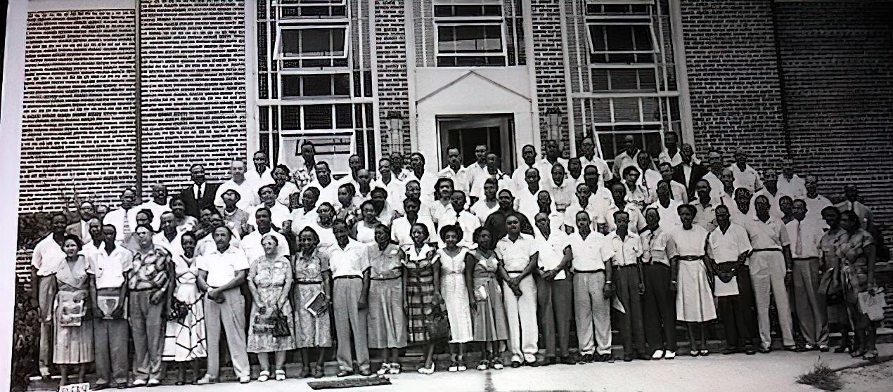 Black and white photo of a group posing in lines in front of the door to a brick school building.