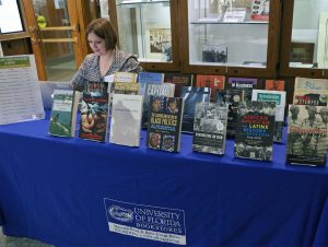 Woman sits behind a table that is covered in a blue University of Florida tablecloth, where three rows of books are on display.