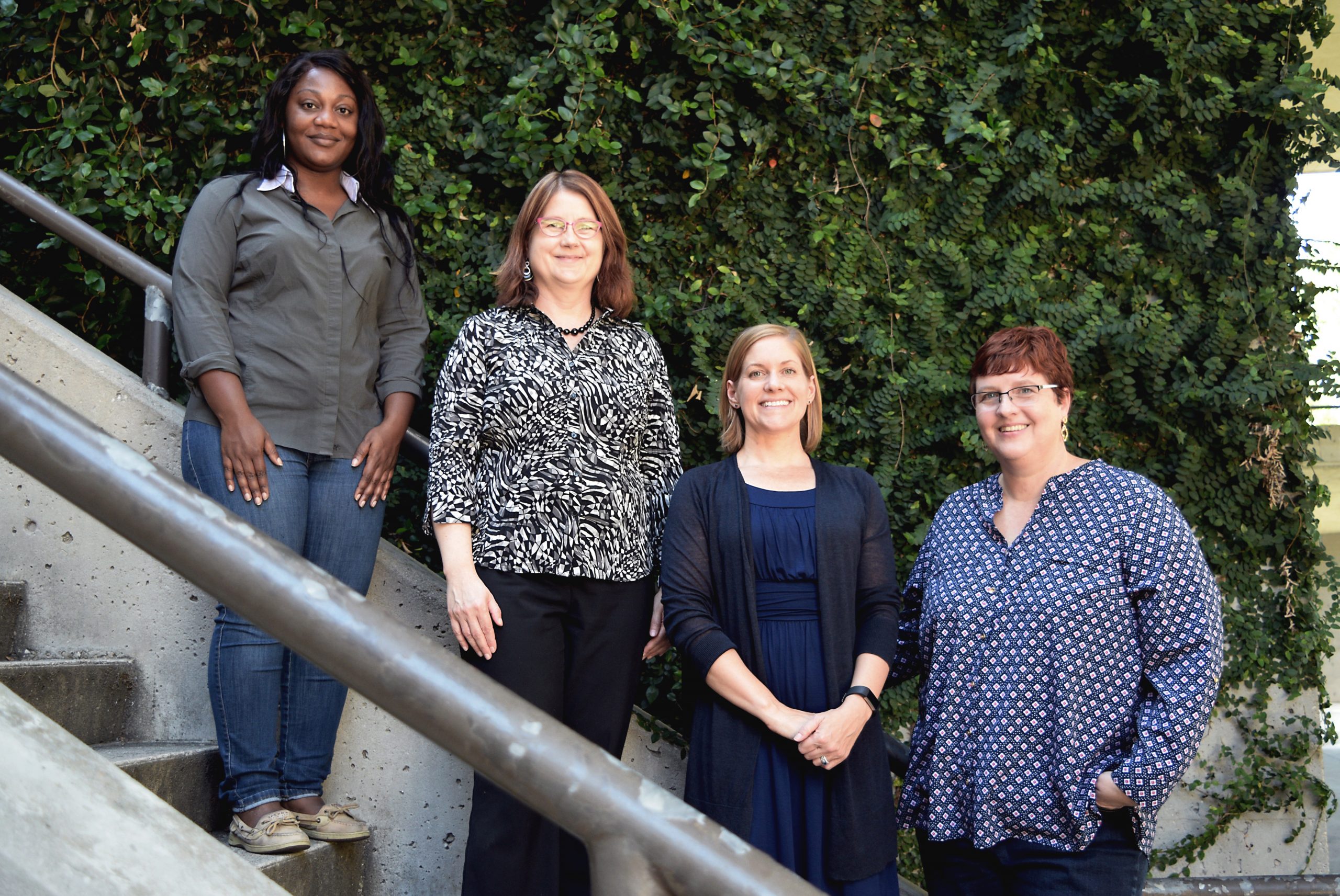 Four women stand on a set of stairs in front of green hedges. They each look at the camera, smiling.