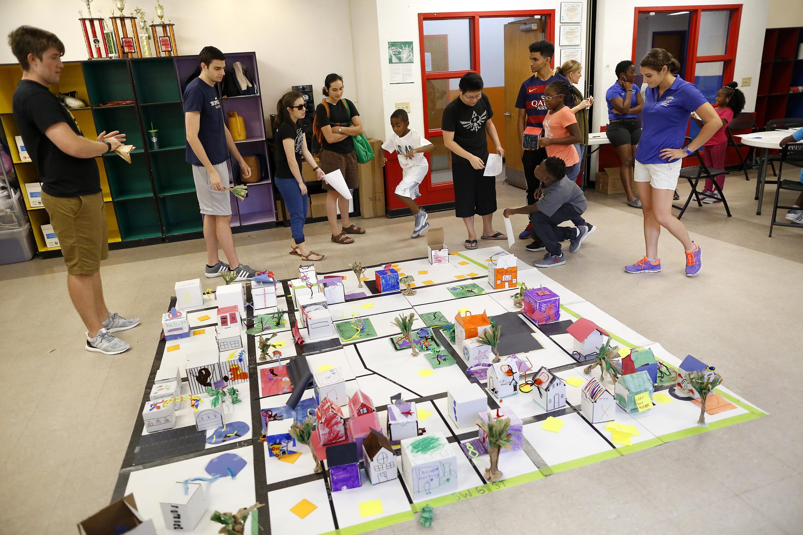 A group of excited students and instructors stand behind a diorama depicting a neighborhood divided by black lines and made of colorful boxes.