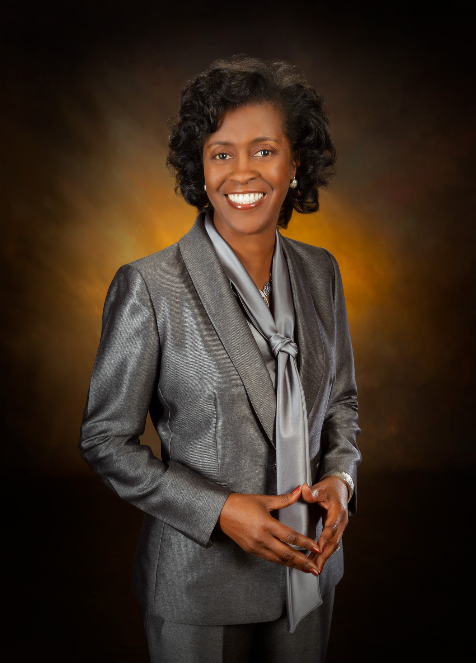 Dr. Adams wears a silver suit and is standing in front of a blurred brown background. She is looking at the camera and smiling.