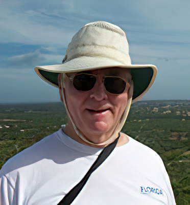 Dr. McCarthy wears a hat, sunglasses, and UF shirt while standing outside in front of a landscape background. He is looking at the camera and smiling.