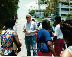 Dr. Baan wears sunglasses. He speaks to a group from the middle of a street, which is edged by cars and tropical plants.