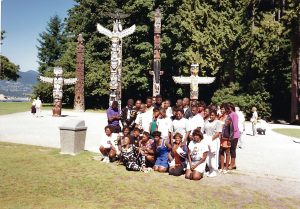 A group poses in front of a series of ornately carved Totem Poles.