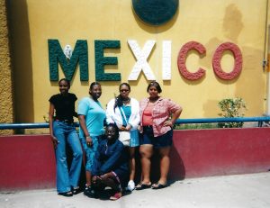 A group poses in front of a wall, where a series of ornate letters spell out Mexico in green, white, and red.