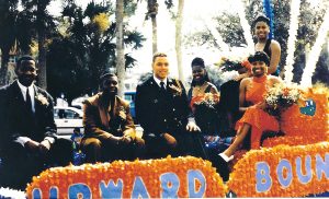 A group poses on a float decorated in orange and blue. Three men wear suits, ties, and corsages; three women wear formal dresses and hold flowers.