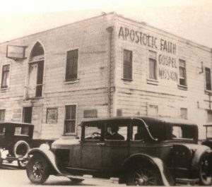 Sepia photo of cars driving past the corner of a building where text has been painted, which reads: Apostolic Faith, Gospel, Mission.