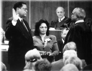 Black and white photo of a courtroom. Stephan stands beside Evelyn with one hand on the Bible and the other raised above his shoulder.