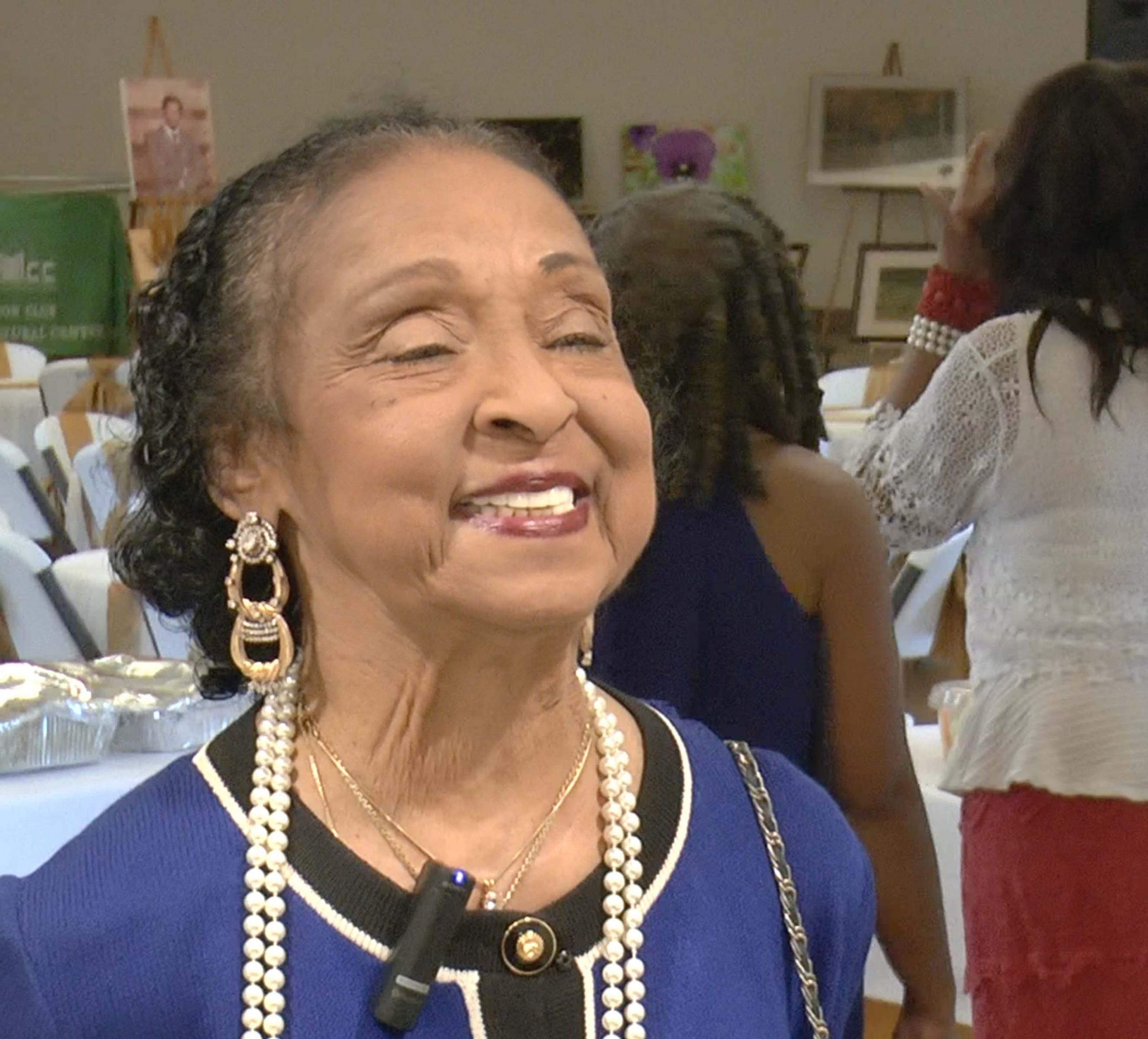 Mrs. Banks wears earrings, pearls, and a blue shirt with black trim. She is smiling, looking up and to the right of the camera.