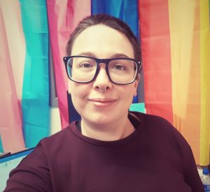 A person with short hair and thick framed glasses standing in front of trans, bisexual, and queer pride flags.