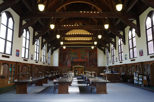 An old reading room with high, wooden ceilings, wooden arches, and two rows of tables with individual lights and chairs