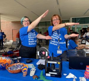 Two people doing a the Gator Chomp standing behind a table. On the table is a 3D printer, laptop, and small 3D printed items