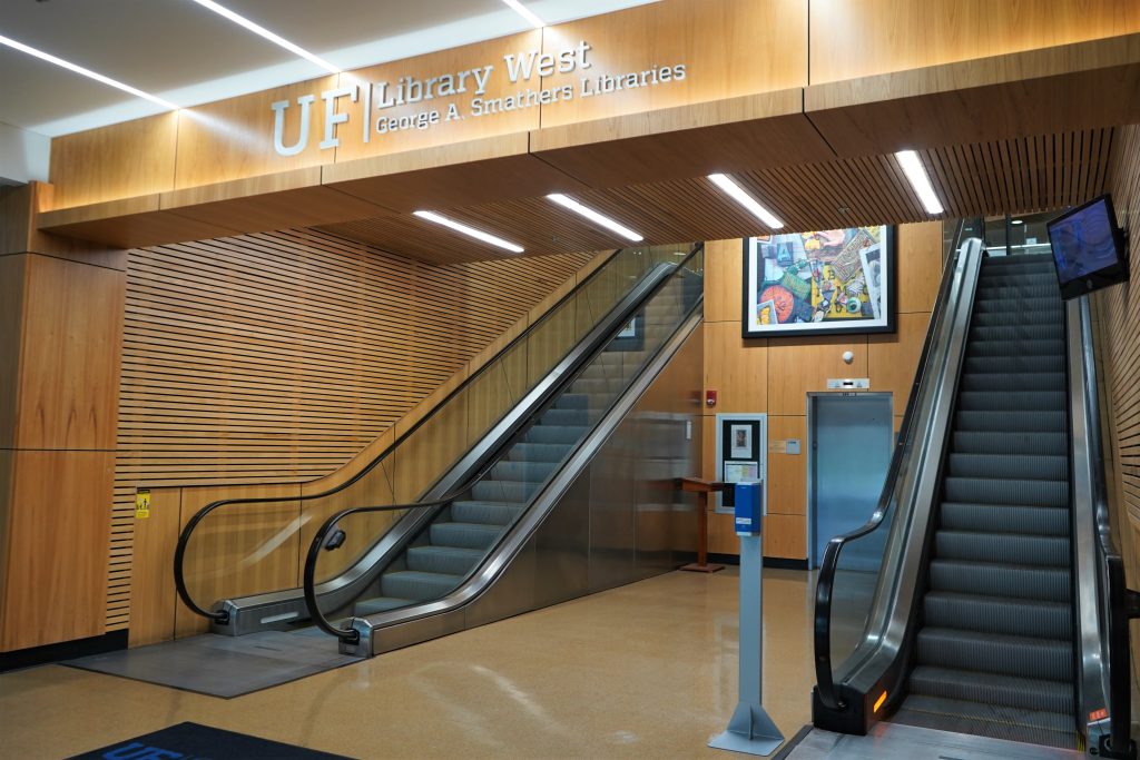 Wood paneled walls and escalators leading upstairs. Overhead, a sign reads "UF Library West, George A. Smathers Libraries"