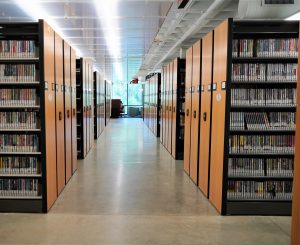 Looking down a walkway lined on either side with library shelving. The closest shelves are full of DVDs