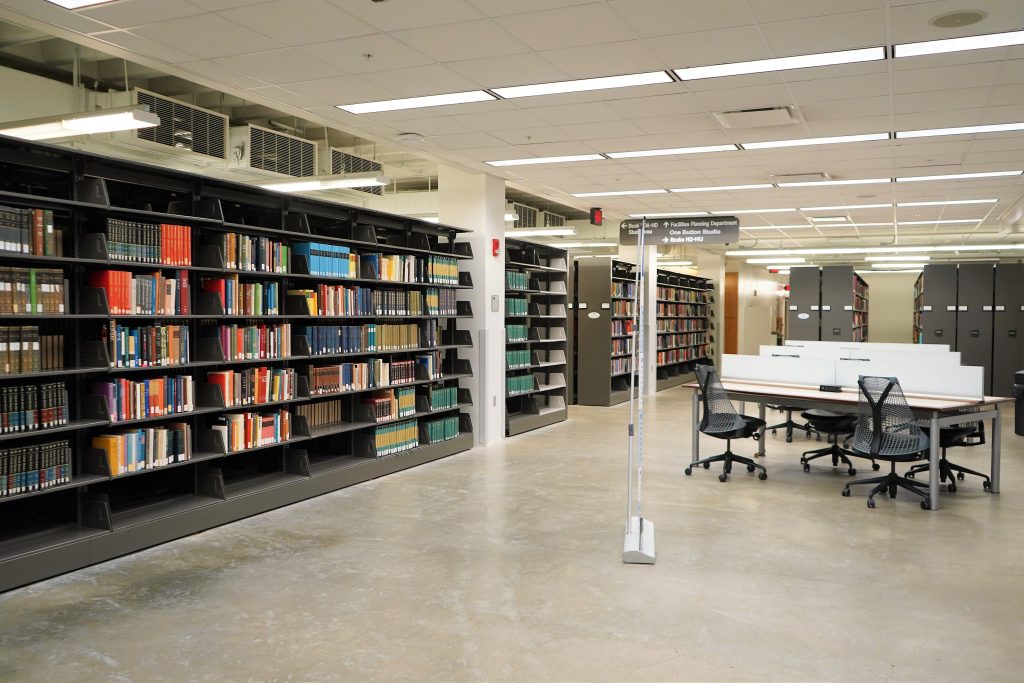 A large room with bright overhead lighting, library shelves, and a few tables with chairs