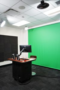 A podium with a monitor. Behind it, a green screen and soundproof foam on the walls. Overhead, bright flood lights.
