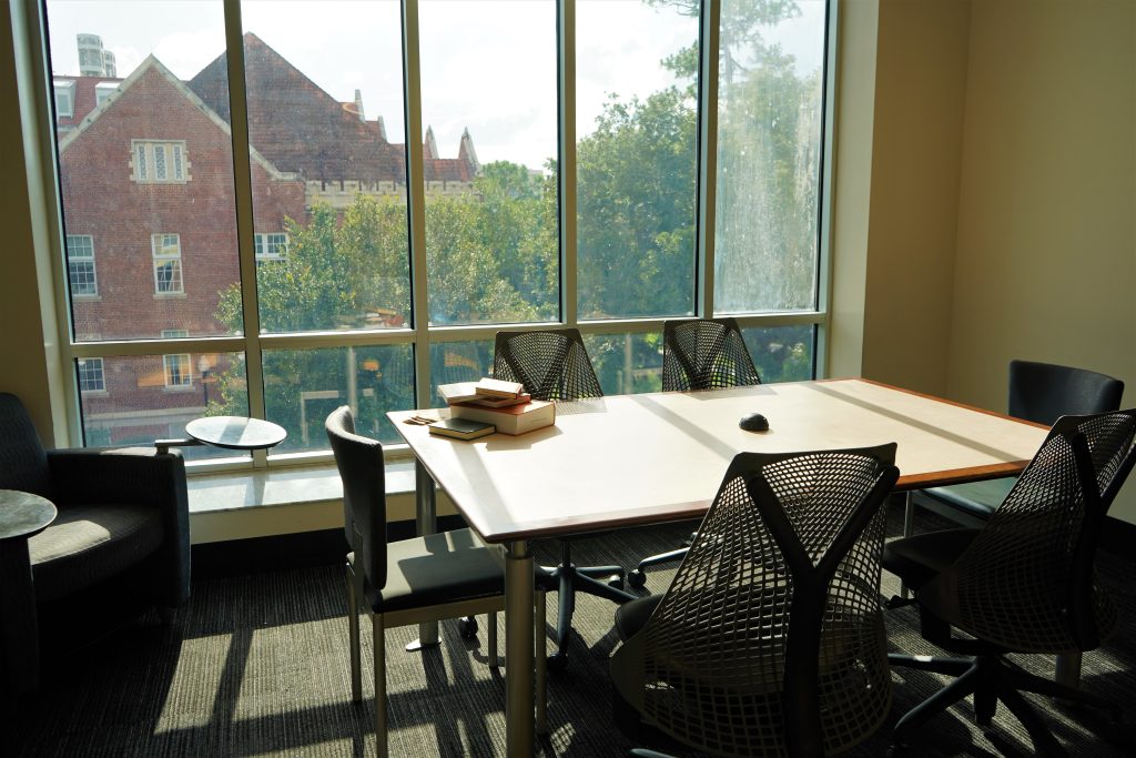 A table with six chairs in front of floor to ceiling windows. The table is bathed in bright sunlight and has a stack of books on top.