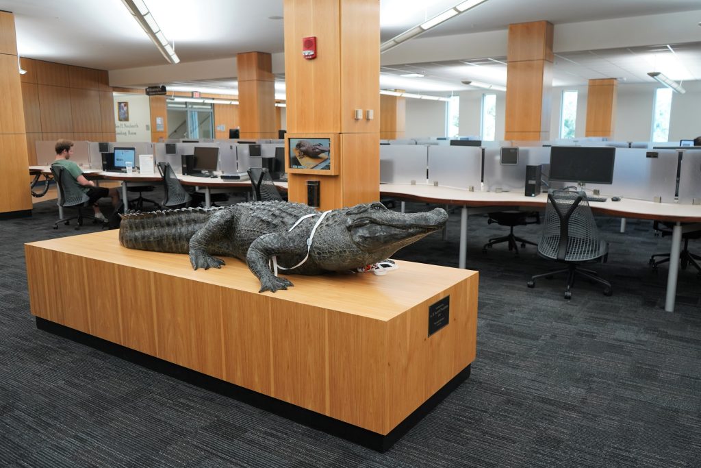 Statue of an alligator on a wooden pedestal. Behind the gator, there are rows of computers and chairs.
