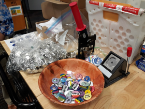 A wooden bowl filled with LGBTQ pride flag buttons, next to a button pressing machine