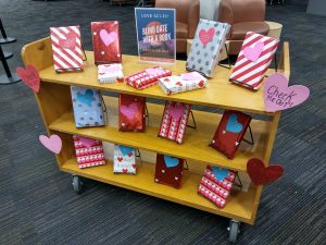 A book cart filled with books in bright, heart-covered wrapping paper to hide the book covers. A sign says "love sci-fi? Check me out! Blind Date with a Book"