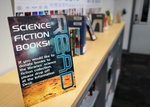 On top of a full bookcase, a sign that says "Read science fiction books! If you would like to donate books to the libraries' science fiction collection, please drop off at the information desk"