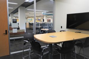 A table with a TV monitor surrounded by chairs. Outside the glass window is the rest of the library floor.