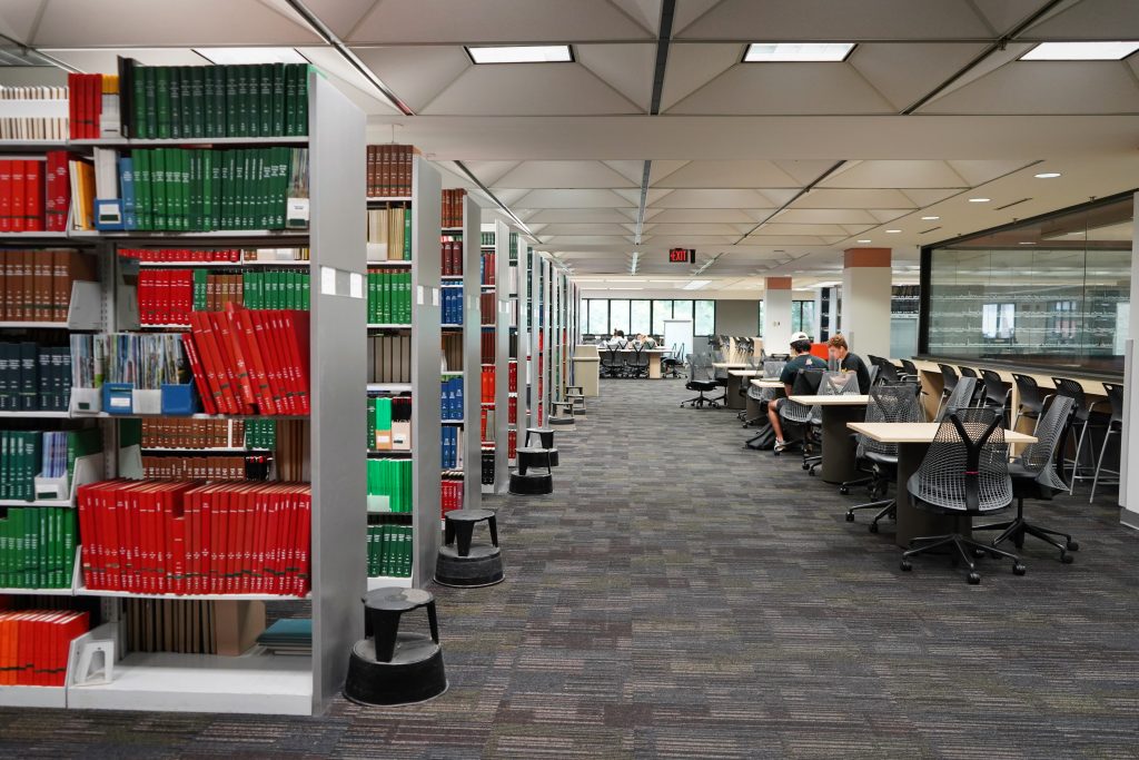 A line of bookshelves with bright, colorful covers. Next to the book stacks and several tables, one of which has students studying
