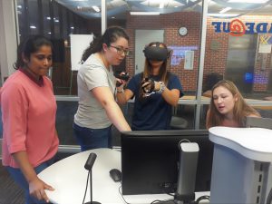 Four students standing behind a computer monitor. One student is wearing a virtual reality headset over their eyes and holding the controllers towards the camera