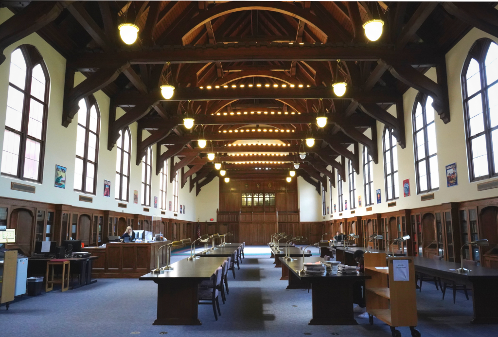 A long room with high vaulted ceilings. The ceiling is adorned with wooden arches. In the room, there are rows of tables with individual desk lamps and seats.