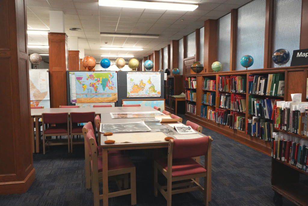 A room lined with wooden bookshelves holding maps and globes. In the center, group study tables with maps laid out to view
