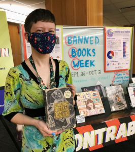 A staff member holding the book The Golden Compass with Library West’s Banned Books Week display in the background