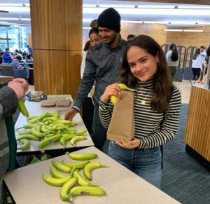 A student in a striped turtleneck shirts poses with a banana and a brown lunch bag in front of a table of bananas