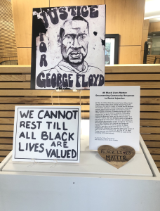 An exhibit case showing protest signs, including one with a hand drawn portrait of George Floyd with the text "Justice for George Floyd" and another with the text "We cannot rest till all Black lives are valued"