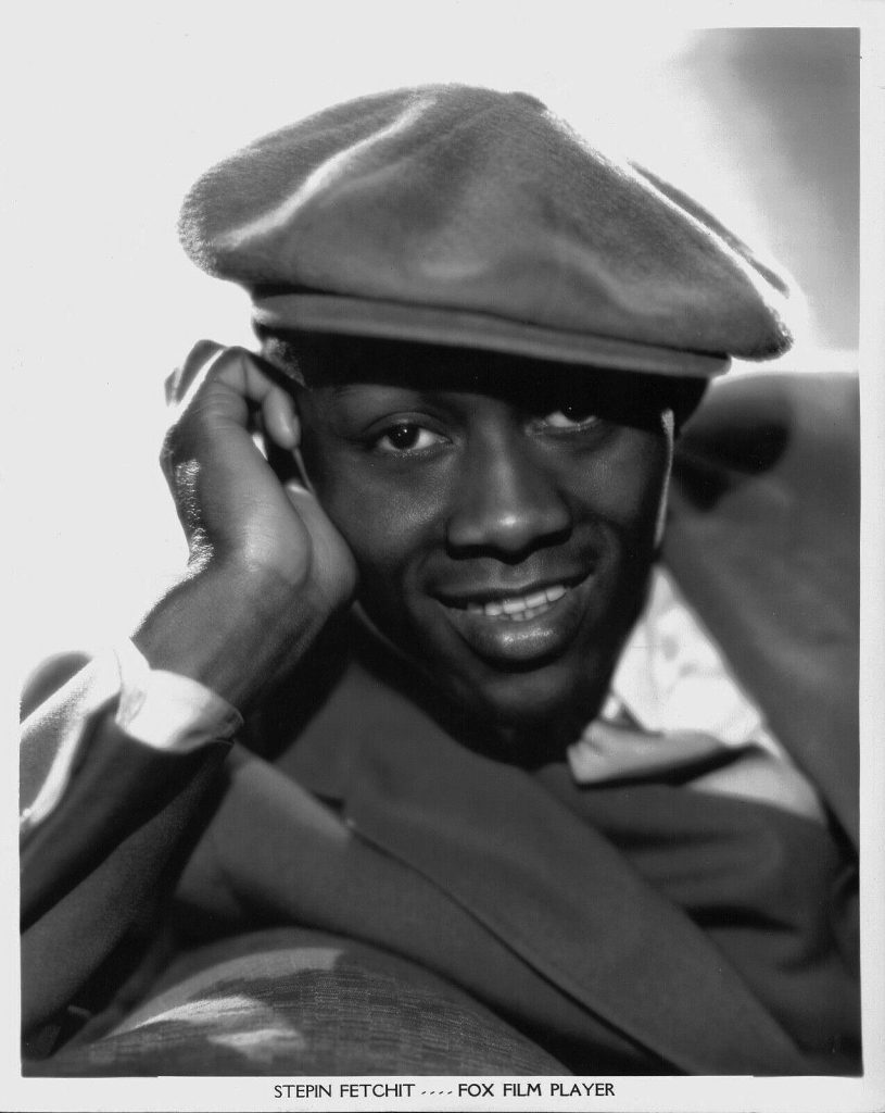 Headshot photo from Fox Film Player, a Black-and-white portrait of a man with the character name, Stepin Fetchit, wearing a flat cap and a suit, smiling and leaning on his right hand.