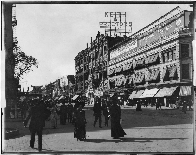 A row of buildings along a street, with people milling about. A large sign that reads "Keith & Proctor's" is above one of the buildings.