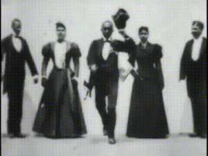 Five dancers, three men and two women, performing a cakewalk. They are all standing, and the man in the middle is holding his top hat off his head.