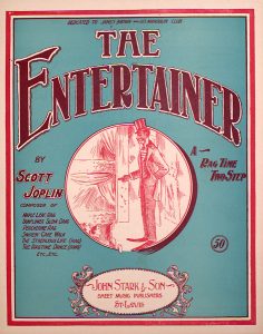 Sheet music cover, with the title "The Entertainer" in large, blocky, curved lettering at the top on a blue background. Under the title is a circular drawing of a Black man in a suit and top hat on a stage, looking out into the audience. Words to the left of the cartoon read "By Scott Joplin, Composer of Maple Leaf Rag, Sunflower Slow Drag, Peacherine Rag, Swipesy Cake Walk, The Strenuous Life (Rag), The Ragtime Dance (Song), Etc., Etc..." To the right of the cartoon are the words "A Rag Time Two Step." Tiny words at the top read "Dedicated to James Brown and His Mandolin Club." At the very bottom is a decorated oval containing the words "John Stark & Son, Sheet Music Publishers, St. Louis."