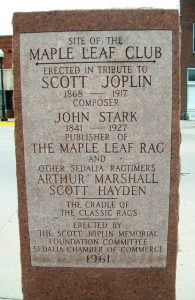 Stone marker with words engraved on it: "Site of the Maple Leaf Club. Erected in Tribute to Scott Joplin, 1868-1917, Composer; John Star, 1841-1927, Publisher of The Maple Leaf Rag; and other Sedalia Ragtimers, Arthur Marshall, Scott Hayden. The Cradle of The Classic Rags. Erected by The Scott Joplin Memorial Foundation Committee, Sedalia Chamber of Commerce, 1961."