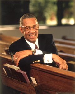 Reverend William Lawson leaning on the back of a church pew bench, smiling.