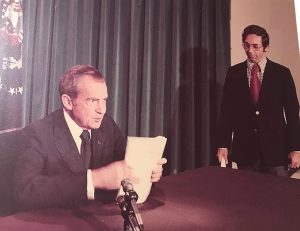 Nixon sitting at a table in front of a microphone, holding papers, preparing to make his resignation speech. White House Deputy Special Assistant Alvin Synder stands to the side of the table.