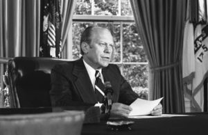 President Gerald Ford sitting at a desk in front of a microphone, holding papers, announcing his decision to pardon former President Richard Nixon. Behind him is a curtained window.