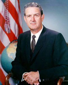 John Connally from the waist up, sitting in a chair and looking at the camera. He is wearing a suit. Behind him is an American flag and a globe.