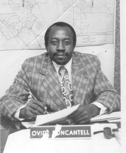 Ovide Duncantell sitting at a desk with paper and pen in hand. On his desk is a nameplate, and behind him on the wall is a map.