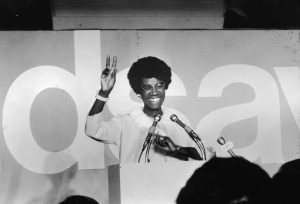 Shirley Chisholm standing at a podium, smiling and waving to a crowd during her acceptance speech for the U.S. House of Representatives.