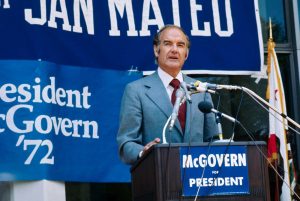 South Dakota senator George McGovern speaking at a podium with microphones during his 1972 presidential campaign. A sign on the podium reads "McGovern for President," and the sign behind him reads "President McGovern '72."