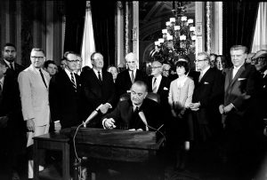 President Lyndon B. Johnson sitting at a table signing the Voting Rights Act of 1965 into law, surrounded by a group of people. The room they are in has large windows with curtains, an ornate painting, and a chandelier. The Voting Rights Act that he is signing prohibits racial discrimination in voting.